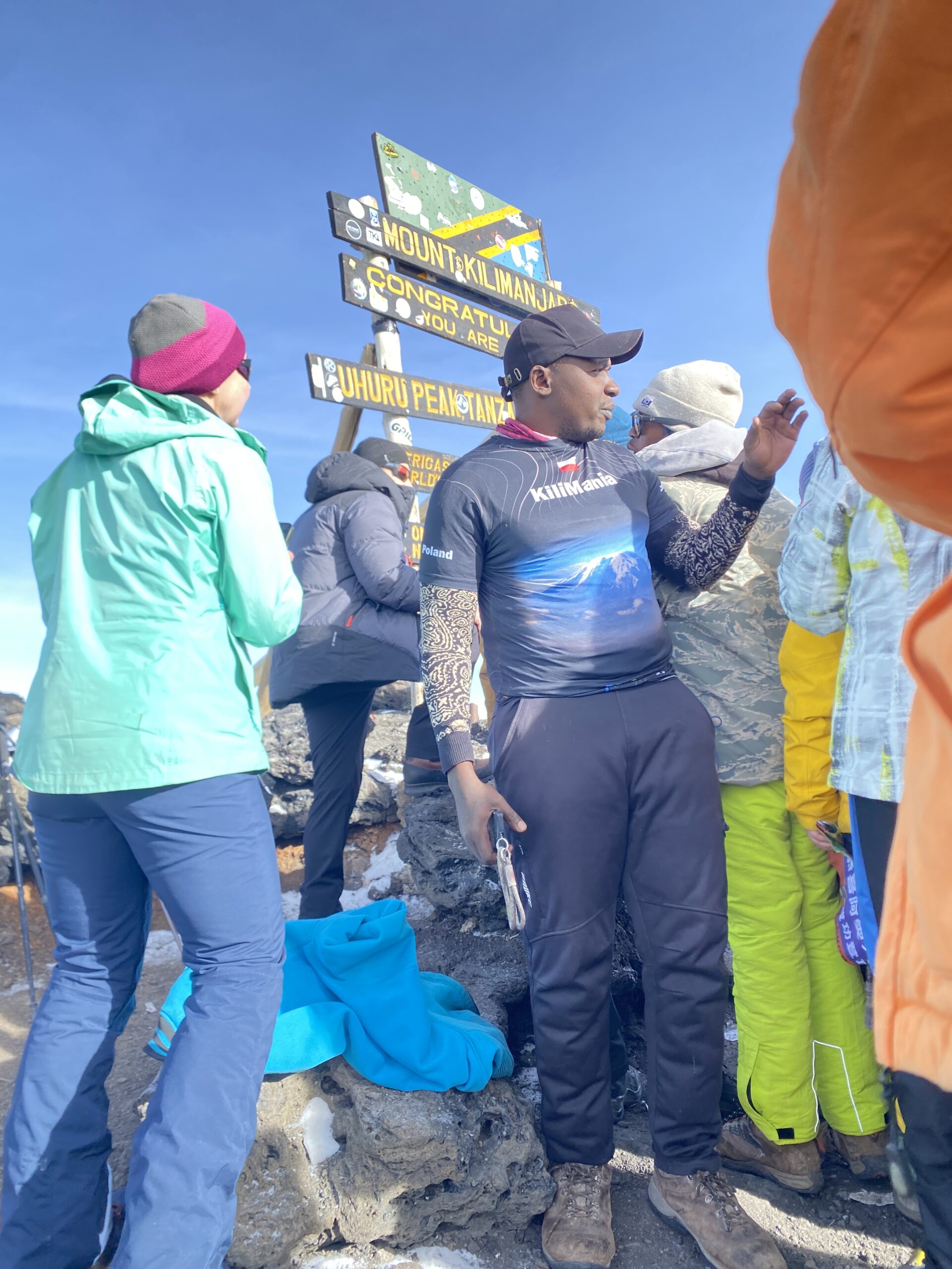 Kilimania Adventure guide Sabinus Msimba at the Mount Kilimanjaro Uhuru Peak summit sign, representing high route success rates in Tanzania. Kilimanjaro routes