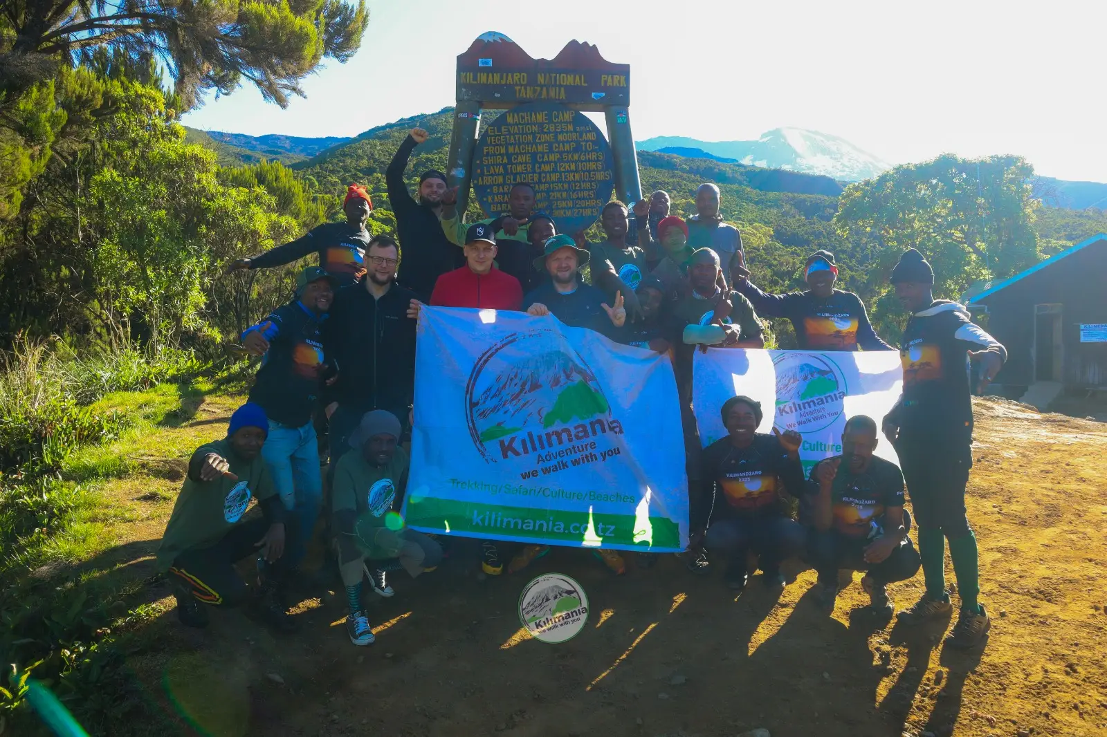 Kilimania Adventure trekking group and guide team posing with company banners at the Machame Camp signpost before heading to Shira Cave Camp, Mount Kilimanjaro. 6 Days Machame Route