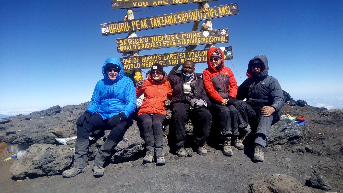 Sabinus Msimba: Senior Kilimanjaro Guide With 22 Years of Experience on Mount Kilimanjaro | Kilimania Adventure Sabinus Msimba, Senior Guide at Kilimania Adventure, seated in the middle of a summit group at the Uhuru Peak sign on Mount Kilimanjaro, Tanzania, Kilimanjaro Success Rate by Route