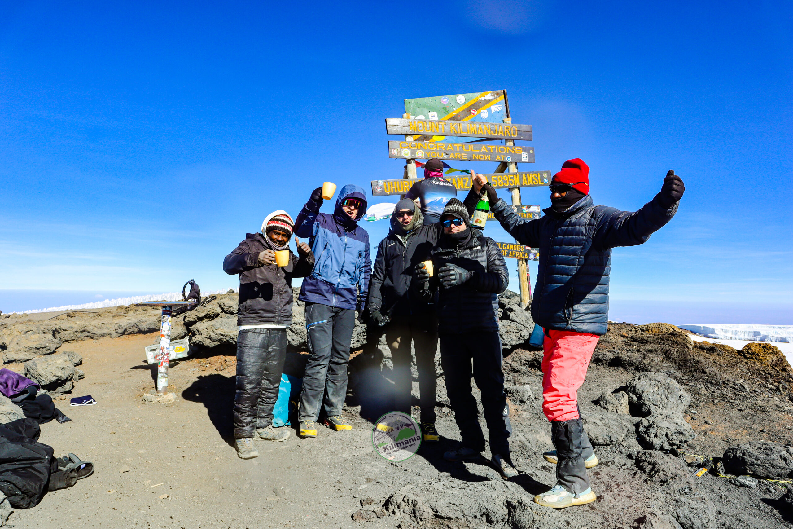 Senior Kilimanjaro guide Sabinus Msimba holding the Uhuru Peak sign with Isack Mlala and climbers after reaching the summit of Mount Kilimanjaro via the Shira Route at 5,895 meters.