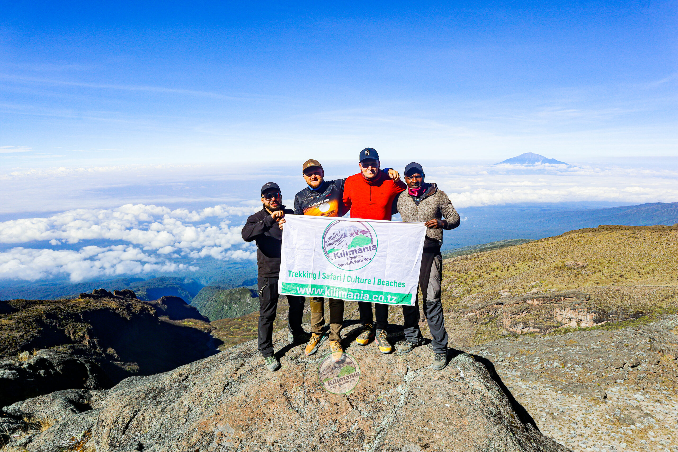 Guide Sabinus Msimba and climbers stand on a scenic viewpoint along the Lemosho Route on Mount Kilimanjaro with Mount Meru visible above the clouds
