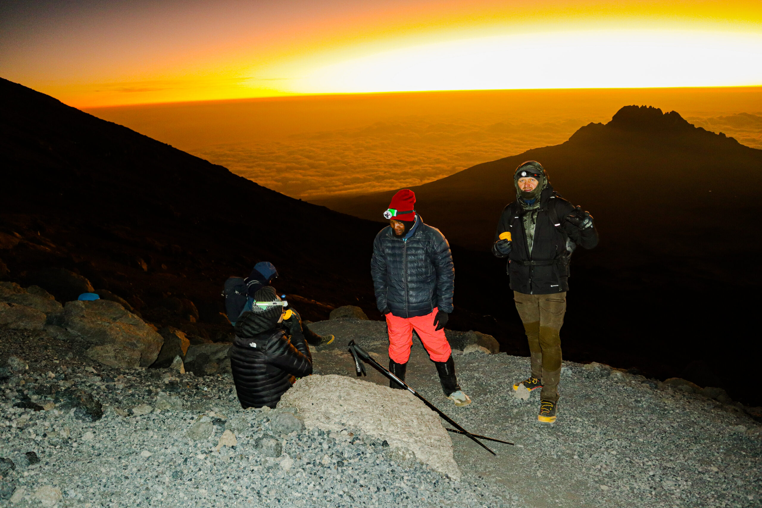 Guide Izack Mlala in red trousers assisting climbers at Stella Point during sunrise on Mount Kilimanjaro — summit support during the final push to Uhuru Peak