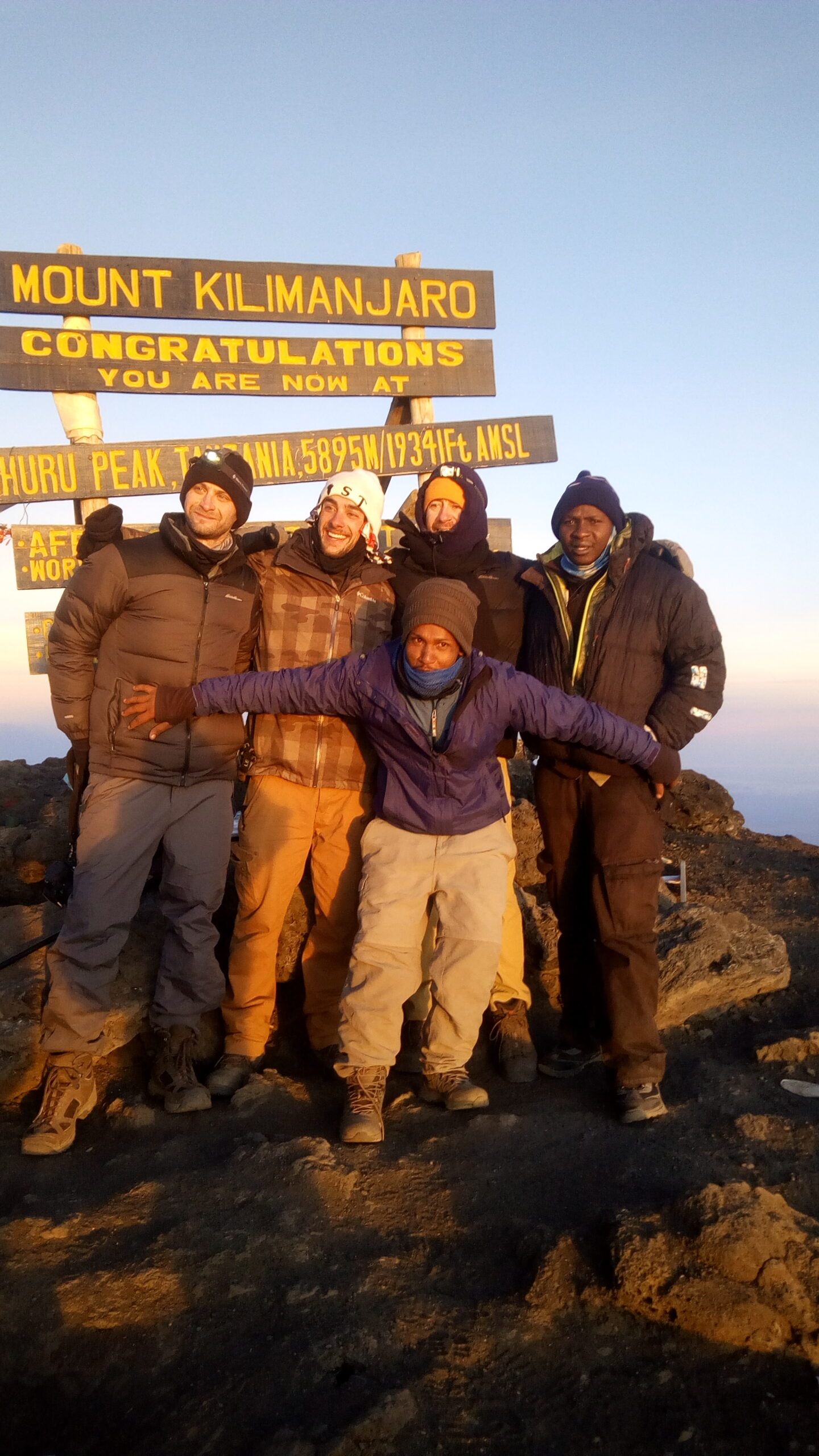 Senior Guide Sabinus Msimba celebrating with Kilimania Adventure guests at Uhuru Peak summit sign on Kilimanjaro