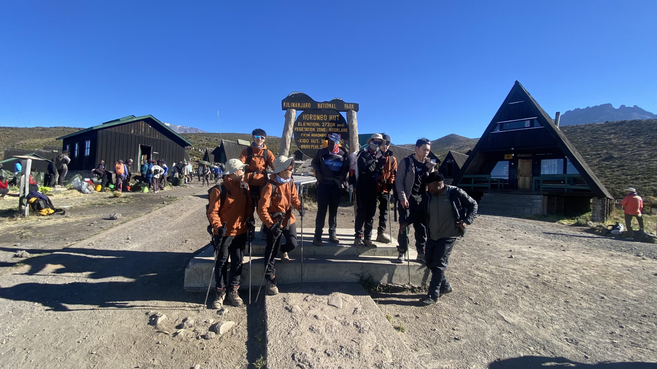 6-Day Marangu Route Trip Report | Kilimania Adventure Eight climbers and their Kilimania guide standing on the concrete steps in front of the yellow wooden Horombo Hut elevation sign, posing with trekking poles under a clear blue sky. 6-Day Marangu Route Trip Report