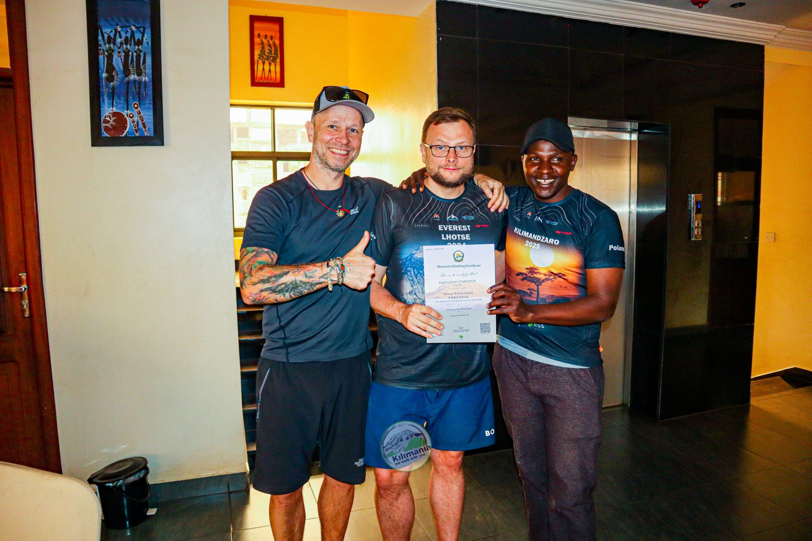 Krzysztof holding a Kilimanjaro summit certificate after reaching Uhuru Peak, with tour leader Szymon and guide Sabinus Msimba