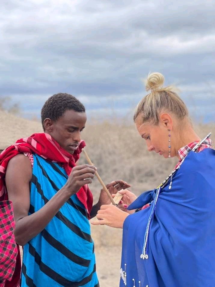 Young Maasai man in a blue and red shuka shows a visitor how to make fire by spinning a wooden stick