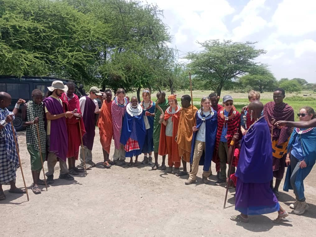 Visitors wearing Maasai shukas and beaded collars pose with their hosts beside a safari vehicle under acacia trees