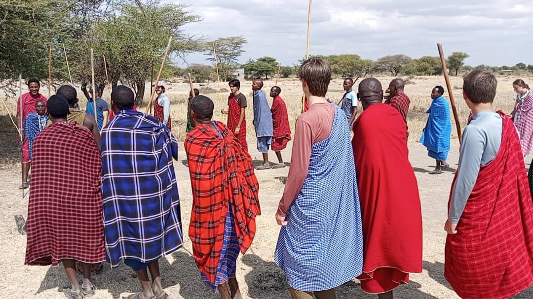 Visitors wrapped in blue and red shukas stand in the welcome circle with Maasai hosts holding staffs