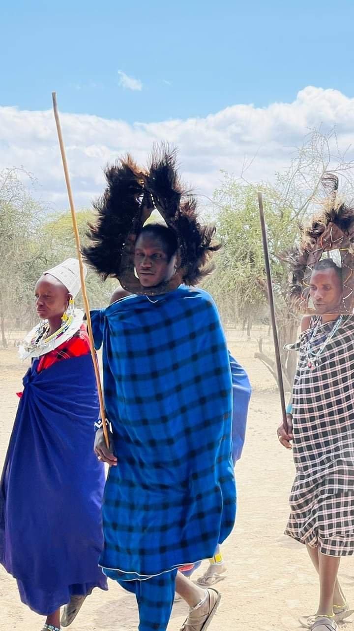 Maasai man in a blue checked shuka wearing a dark feather headdress, walking beside a woman in beaded collar